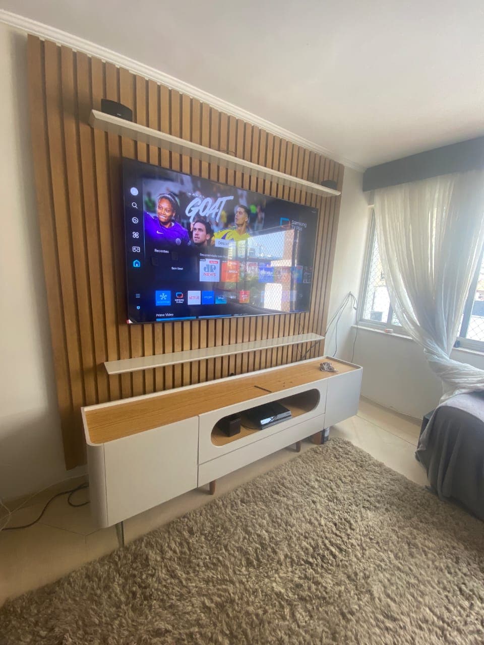 Painel ripado de madeira ocupando a parede da sala, com TV instalada e rack branco de pés palito