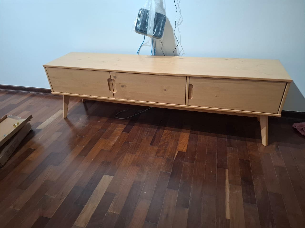 Rack de TV em madeira clara com três portas e pés palito, montado sobre piso de madeira escura